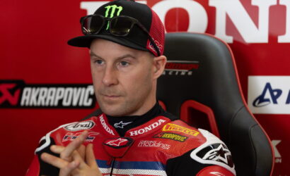Motorcycle racer in a red Honda suit sits in a chair during an interview, wearing a Monster cap and sunglasses on top of the cap.