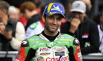 Motocross/MotoGP rider in green Castrol racing suit smiles at the camera, wearing a blue Michelin cap, with blurred fans in the background.