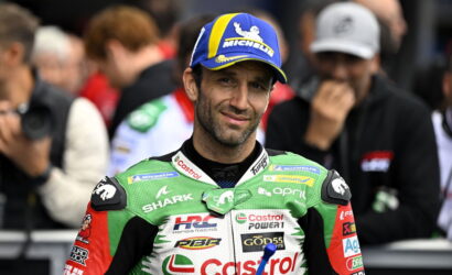 Motocross/MotoGP rider in green Castrol racing suit smiles at the camera, wearing a blue Michelin cap, with blurred fans in the background.
