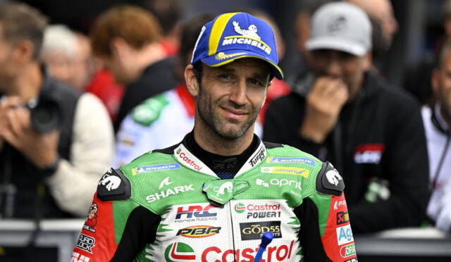 Motocross/MotoGP rider in green Castrol racing suit smiles at the camera, wearing a blue Michelin cap, with blurred fans in the background.
