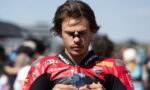 Young male motorcycle racer in red and black leather gear examines a small blue prize or trophy in his hands at a race event, crowd blurred in the background.