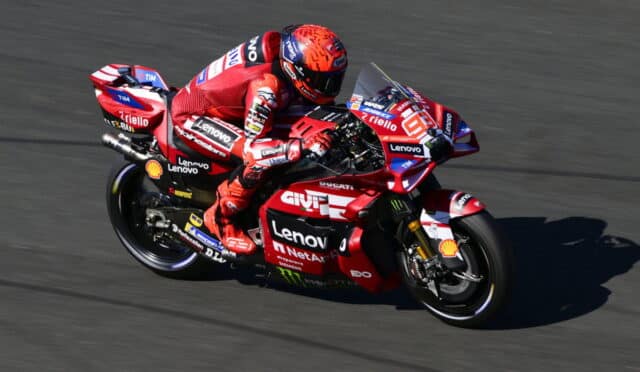 Red Ducati MotoGP bike in a sharp left turn, rider in red racing suit leaning into the corner with sponsor logos all along the fairing and bodywork of the bike.