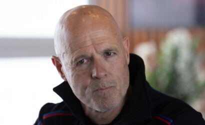 Close-up of a bald man with a contemplative expression, wearing a dark jacket with red and blue stripes.