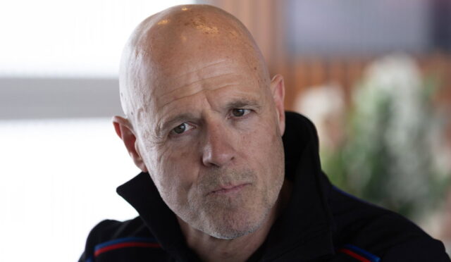 Close-up of a bald man with a contemplative expression, wearing a dark jacket with red and blue stripes.