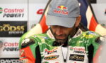 Racing driver in a Red Bull cap and green racing suit looking down, with sponsor banners behind him.