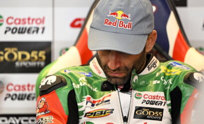 Racing driver in a Red Bull cap and green racing suit looking down, with sponsor banners behind him.