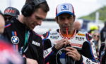 Motorsport rider in a ROKIT racing suit wearing a cap, sipping from a drink tube while a teammate watches nearby.