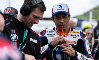 Motorsport rider in a ROKIT racing suit wearing a cap, sipping from a drink tube while a teammate watches nearby.