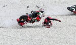 Motorcycle race crash on a gravel track: rider slides while another rider tips over nearby, debris flying around.
