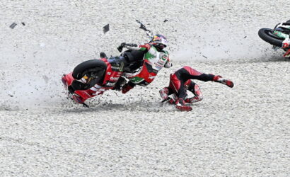 Motorcycle race crash on a gravel track: rider slides while another rider tips over nearby, debris flying around.