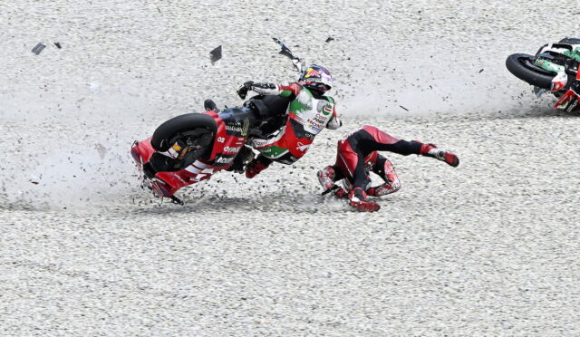 Motorcycle race crash on a gravel track: rider slides while another rider tips over nearby, debris flying around.