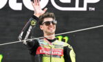Smiling motorcycle racer in a bright yellow and black Enduro suit waves from a podium, wearing sunglasses.