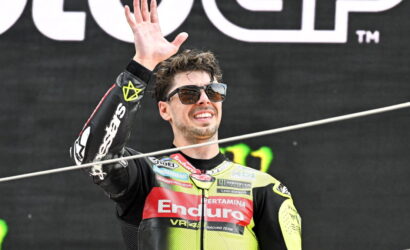 Smiling motorcycle racer in a bright yellow and black Enduro suit waves from a podium, wearing sunglasses.