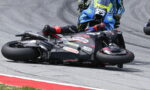 Motorcycle racer slides on the track during a corner, another blue bike close behind on the racing line.