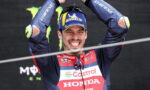 Male motorcycle racer in a blue-red Honda Castrol suit raises arms in victory on a podium, smiling broadly.