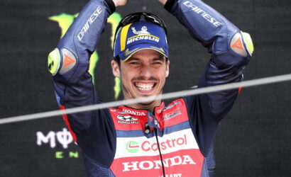 Male motorcycle racer in a blue-red Honda Castrol suit raises arms in victory on a podium, smiling broadly.