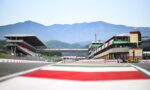 Race track at Mugello Circuit with grandstands and pit complex, mountains in background.