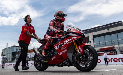 Motogp rider in red racing suit and helmet sits on a red race bike as a team member pushes from behind in the pit lane on a sunny day.