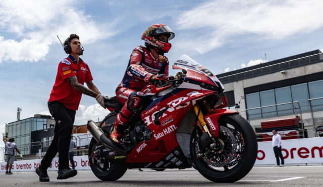 Motogp rider in red racing suit and helmet sits on a red race bike as a team member pushes from behind in the pit lane on a sunny day.