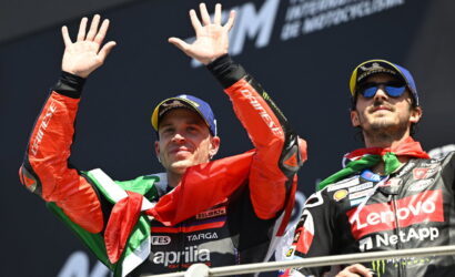 Two MotoGP riders on a podium celebrate, one waving with hands up and an Italian flag draped over his shoulders.