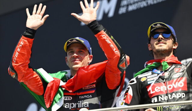 Two MotoGP riders on a podium celebrate, one waving with hands up and an Italian flag draped over his shoulders.