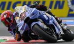 Motorcycle racer in blue-and-white bike leaning hard into a corner on a race track with red and white curbs and a yellow sponsor barrier in the background.