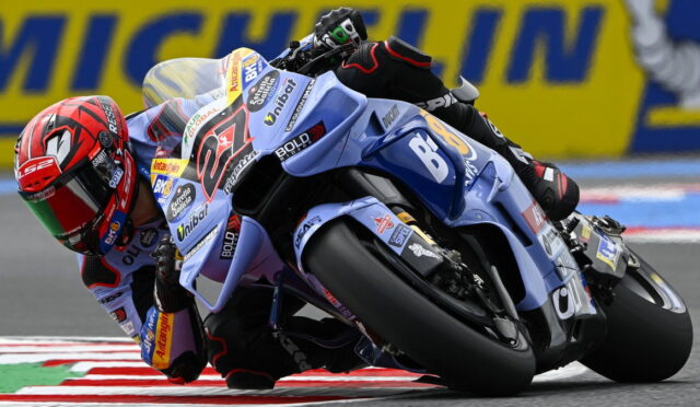 Motorcycle racer in blue-and-white bike leaning hard into a corner on a race track with red and white curbs and a yellow sponsor barrier in the background.