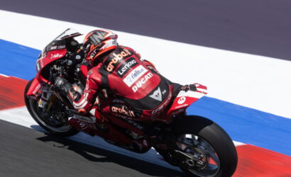 MotoGP rider in red Ducati racing suit leans into a track turn on a red Ducati bike with sponsor logos visible on the suit and bike, walled by blue and red curbing.