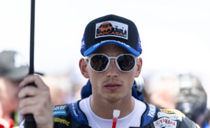 Portrait of a motorbike racer facing the camera, wearing a blue cap with a Vap Casa logo and white sunglasses in racing gear.