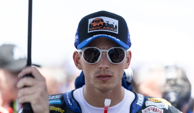 Portrait of a motorbike racer facing the camera, wearing a blue cap with a Vap Casa logo and white sunglasses in racing gear.