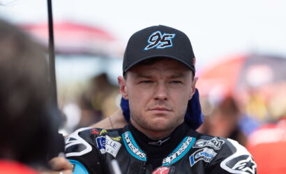 Male motorcycle racer in a black/blue suit and cap with a '95' logo, looking focused toward the left. Image shows his racing gear and sponsor patches close up in a pit area.