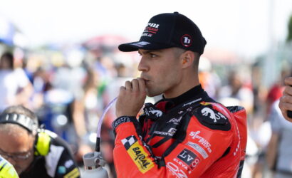 Race car driver in a red racing suit sipping through a straw from a water bottle at a busy pit area, wearing a black cap.