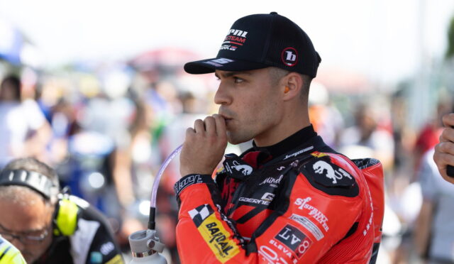 Race car driver in a red racing suit sipping through a straw from a water bottle at a busy pit area, wearing a black cap.