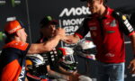 Two motorbike racers shake hands on a podium, fans in the background and racing logos visible behind them (RACINE and Monster energy).
