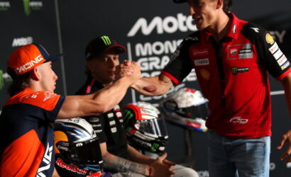 Two motorbike racers shake hands on a podium, fans in the background and racing logos visible behind them (RACINE and Monster energy).