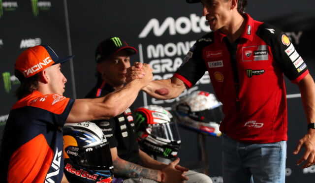 Two motorbike racers shake hands on a podium, fans in the background and racing logos visible behind them (RACINE and Monster energy).