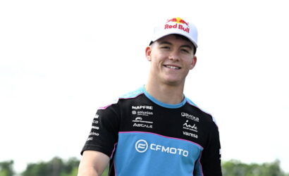 Smiling young male cyclist wearing a blue and black CF Moto team jersey and a white Red Bull cap outdoors.