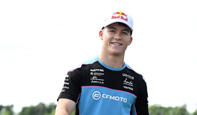 Smiling young male cyclist wearing a blue and black CF Moto team jersey and a white Red Bull cap outdoors.