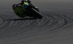 Motorbike racer in neon green gear leans into a sharp turn on a dark race track, leaving curved tire marks behind.