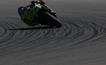 Motorbike racer in neon green gear leans into a sharp turn on a dark race track, leaving curved tire marks behind.