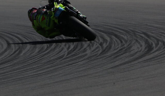 Motorbike racer in neon green gear leans into a sharp turn on a dark race track, leaving curved tire marks behind.