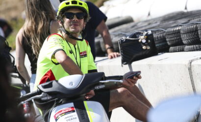 Man in a neon yellow helmet and jersey sits on a scooter, wearing sunglasses and holding a smartphone beside a concrete barrier and stacked tires.