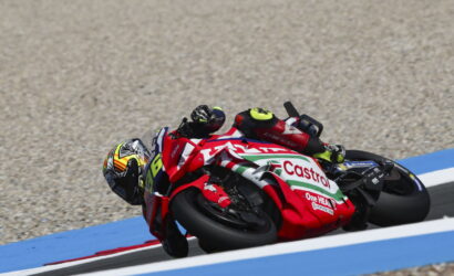 Motorcycle racer on a red-and-white Castrol bike leans into a sharp turn on a blue-curbed racetrack with gravel run-off nearby.