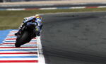 Motorcycle racer leans into a tight corner on a racetrack, riding along red-white-blue striped curbs.
