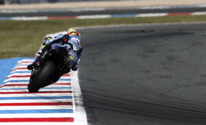 Motorcycle racer leans into a tight corner on a racetrack, riding along red-white-blue striped curbs.