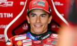 Smiling male motorcycle racer in a red suit and cap with sponsor logos visible on his gear and background.