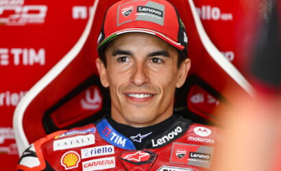 Smiling male motorcycle racer in a red suit and cap with sponsor logos visible on his gear and background.