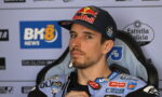 Male motorbike racer wearing a Red Bull cap and blue sponsor-labeled racing suit, seated in a press backdrop.
