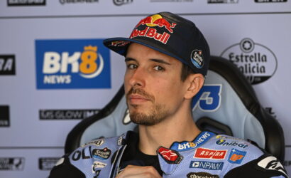Male motorbike racer wearing a Red Bull cap and blue sponsor-labeled racing suit, seated in a press backdrop.
