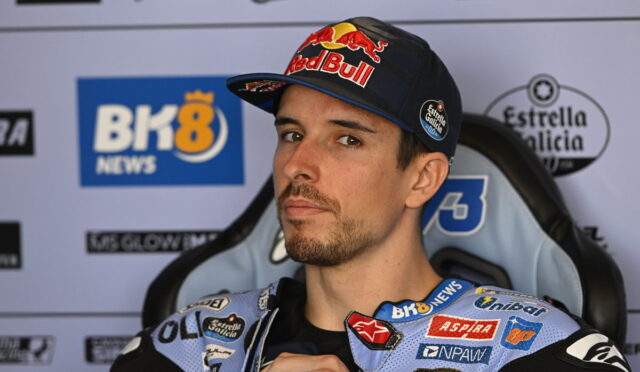 Male motorbike racer wearing a Red Bull cap and blue sponsor-labeled racing suit, seated in a press backdrop.